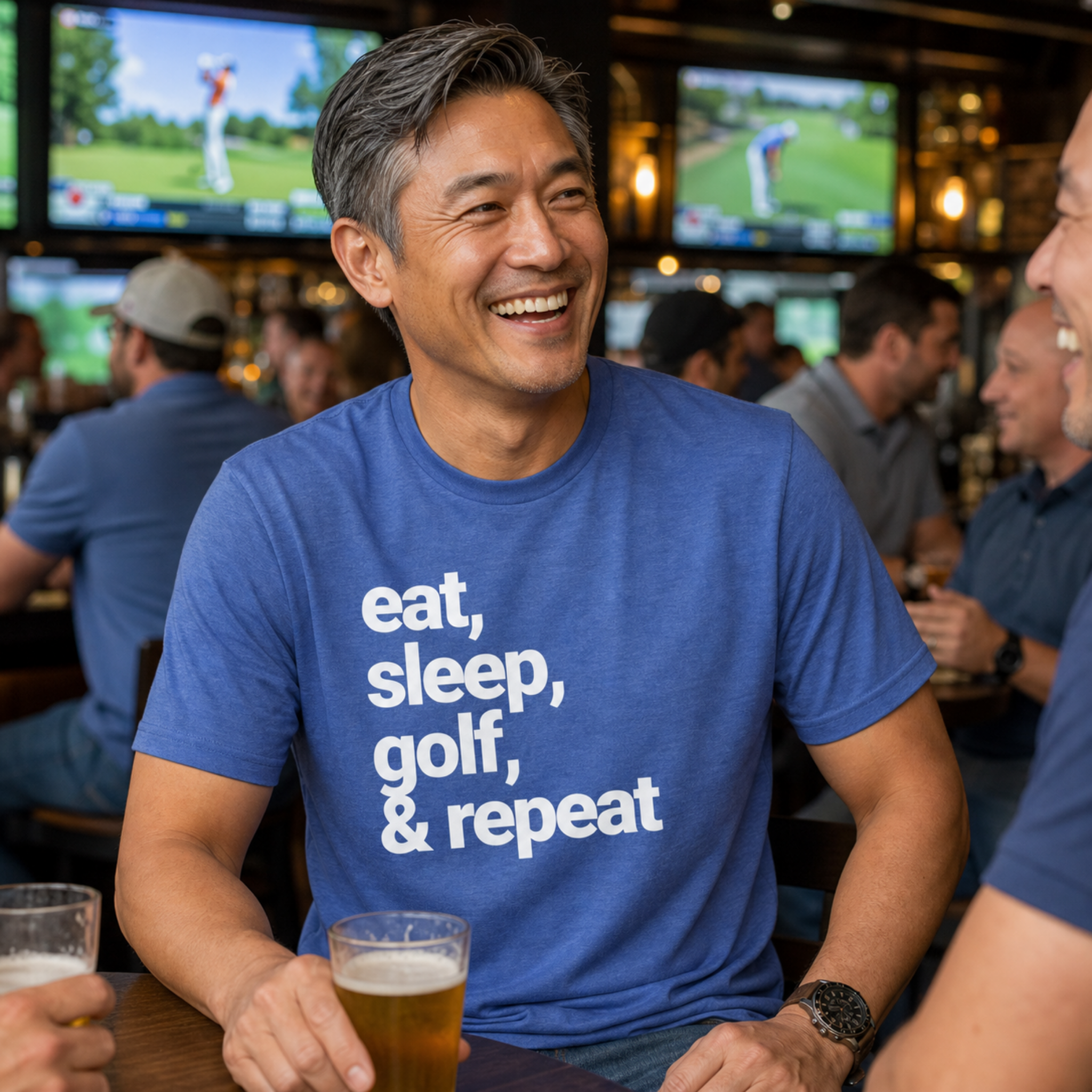 A smiling man in a bar wearing a blue graphic t-shirt with the white text 'eat, sleep, golf, & repeat'. He is holding a glass of beer, with other people and TV screens visible in the background.